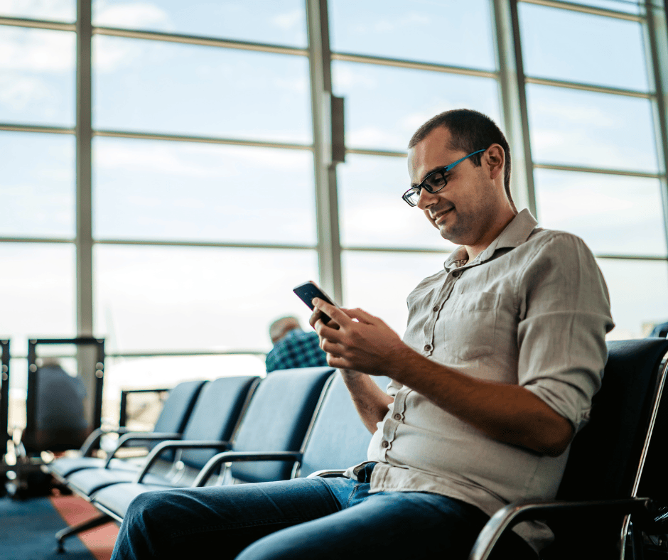 A man with glasses sitting in an airport terminal using his smartphone.