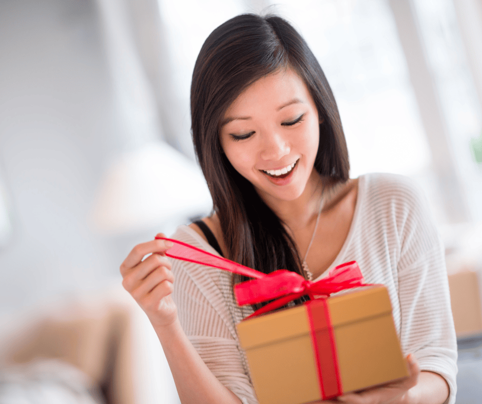 A smiling woman opening a gold gift box with a bright red ribbon in a bright, indoor setting.