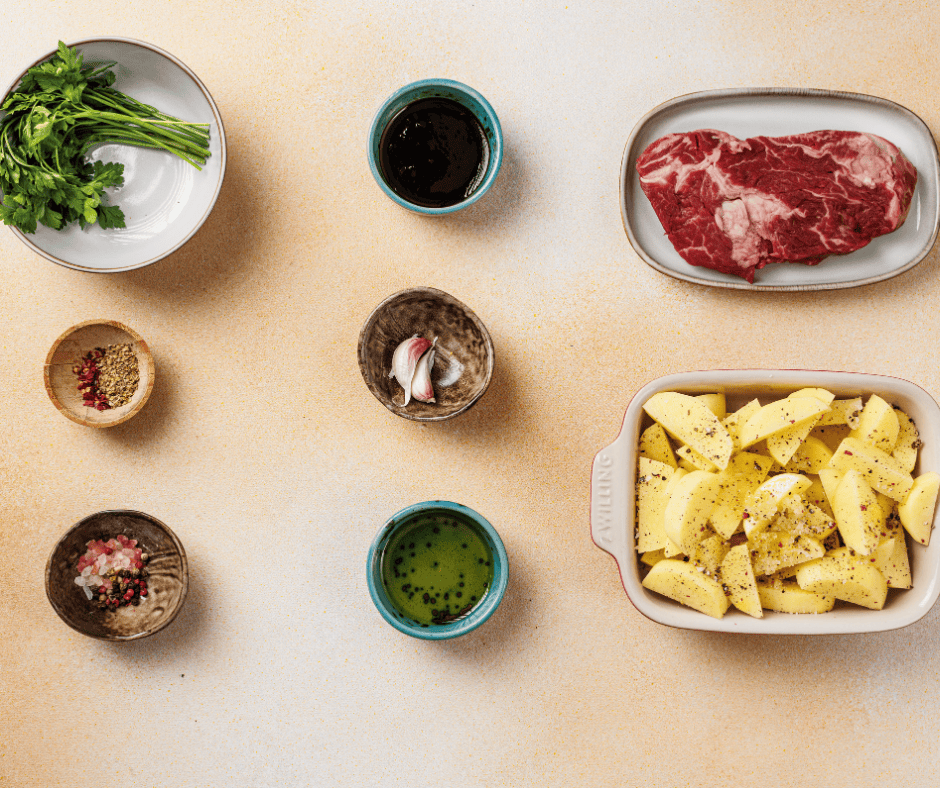 Raw ribeye steak, sliced potatoes, fresh parsley, garlic cloves, and red pepper flakes arranged for meal prep.