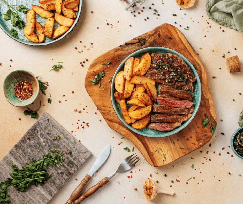 A complete dish of sliced grilled steak with green chimichurri sauce and crispy garlic potato wedges on a wooden board.