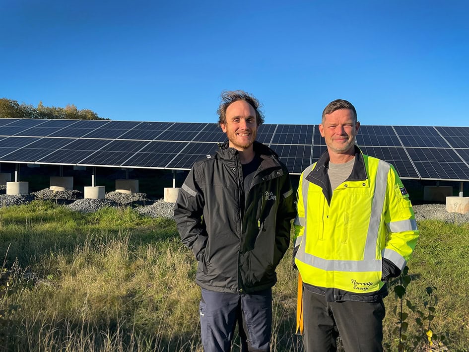 Frode och Tobias står med solelparken bakom sig, de ser in i kameran och ser nöjda ut. Utomhus med blå bakgrund och grönt gräs. 