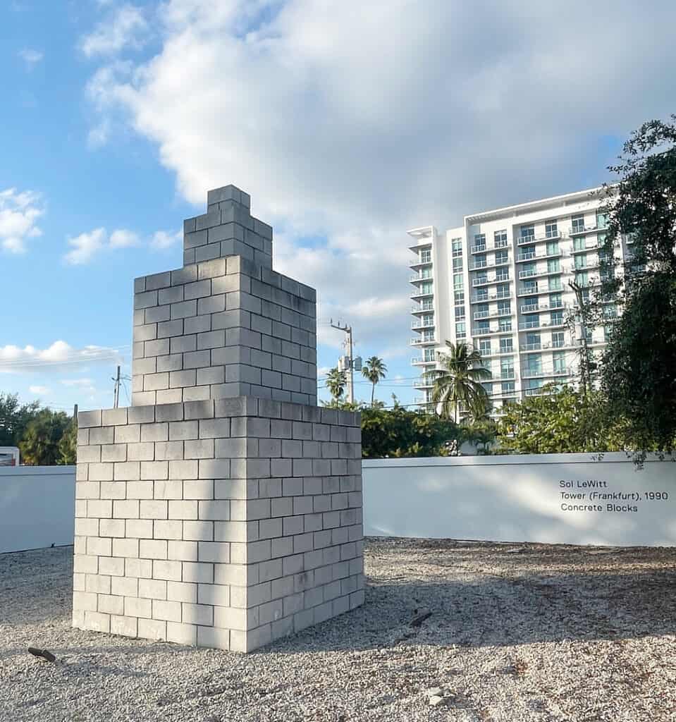 A geometric outdoor sculpture made of stacked concrete blocks stands on gravel, with a modern white apartment building, trees, and a cloudy blue sky in the background. A small wall plaque marks its presence at Miami Art Week 2025.