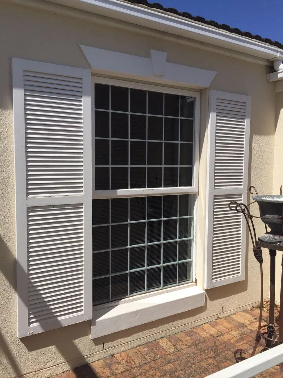 White cottage shutters framing slimline cottage-pane window bars on a cream stucco house under a clear blue sky.
