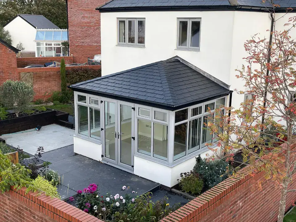 White conservatory with a dark tiled roof beside a white house and paved garden