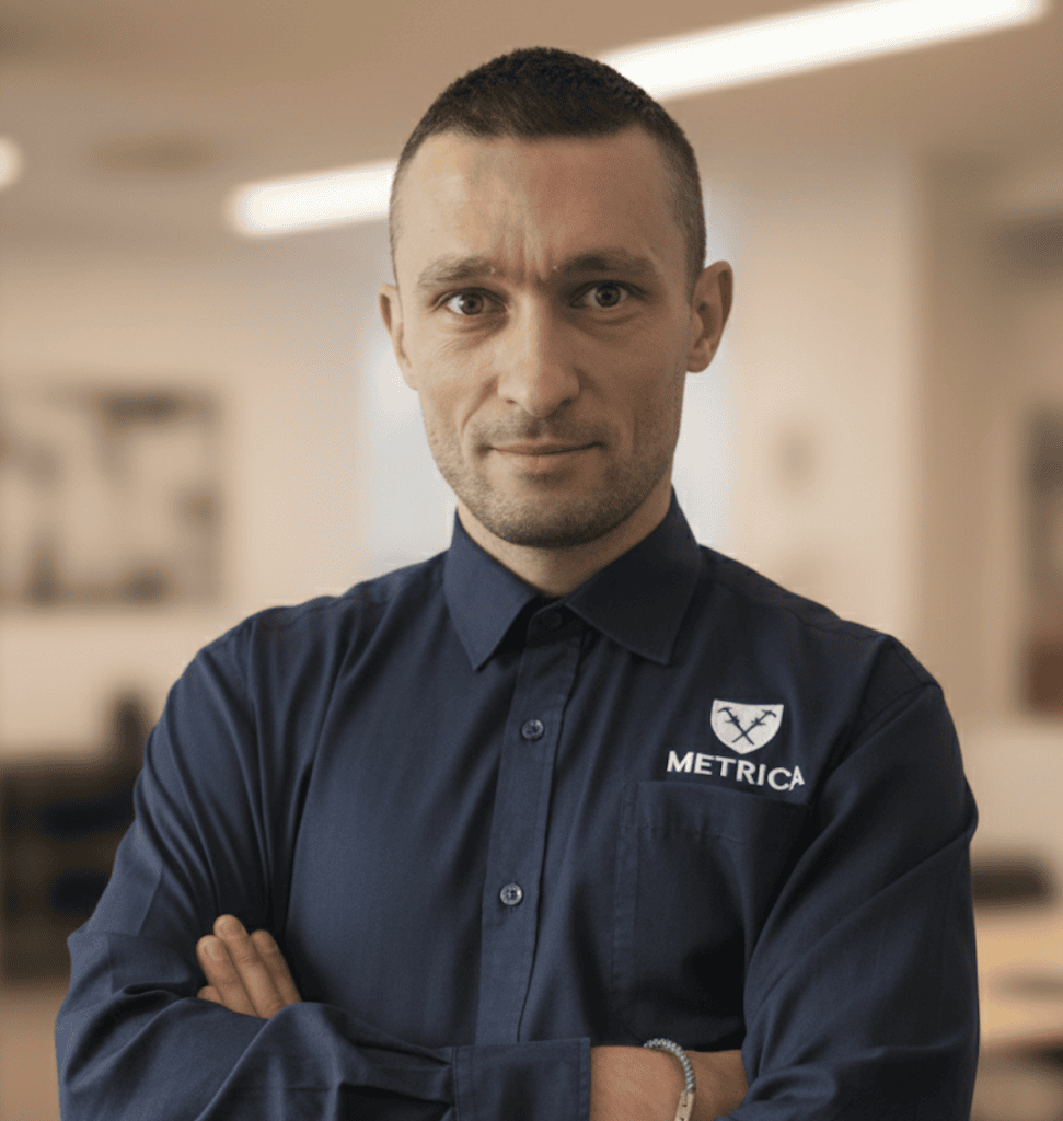 Professional man with short dark hair in a navy button-down shirt bearing the METRICA logo, arms crossed in a blurred office setting
