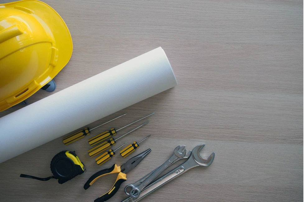 Tools like hard hat, tape, screwdrivers, pliers, wrenches, and plans lie on wooden surface.