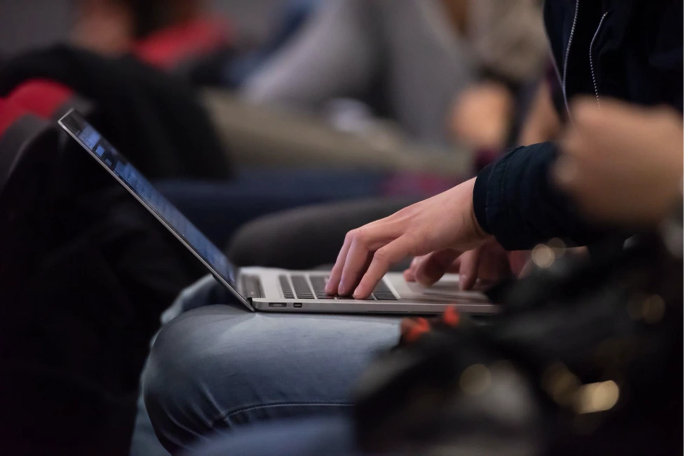 Person types on laptop in casual setting, hands focused; others blurred, suggesting work or study.