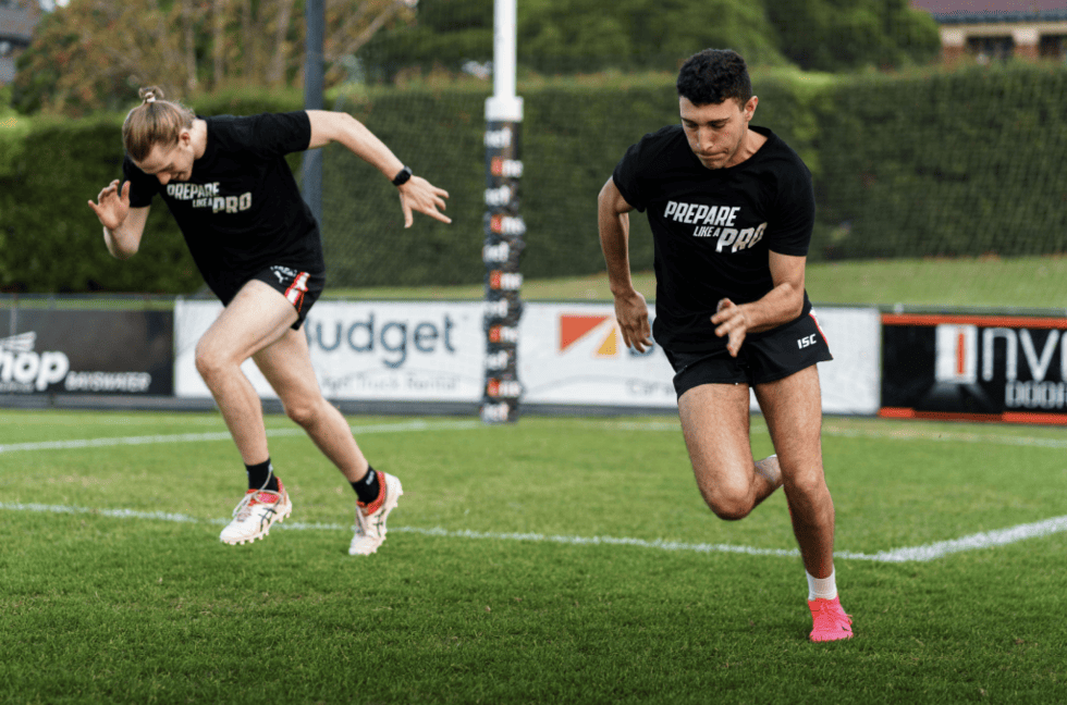 Two athletes sprinting on a field, wearing black shirts with the text 'PREPARE LIKE A PRO', showcasing their speed and focus during training.