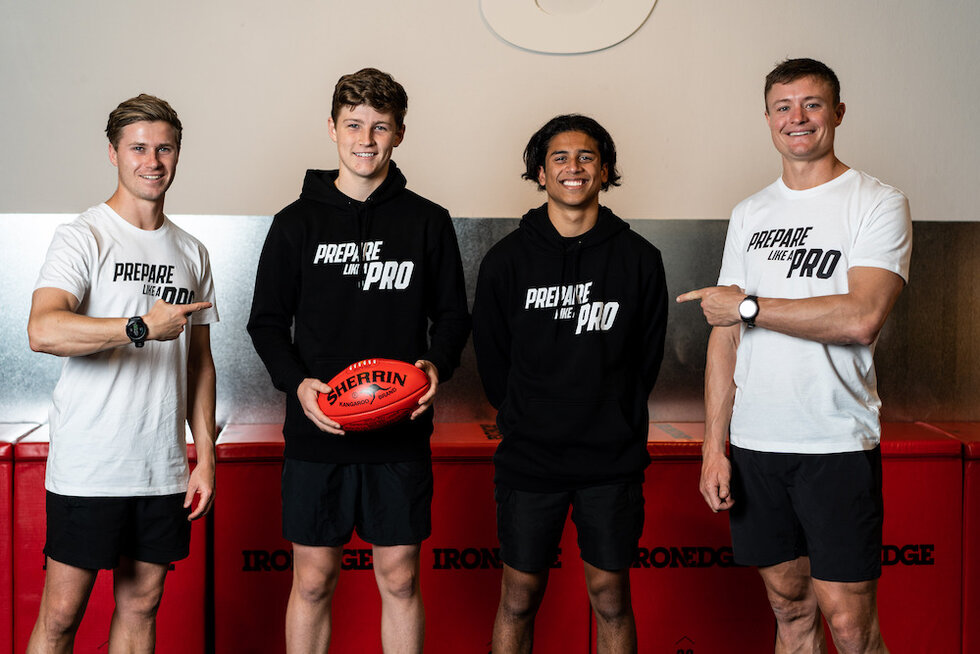 Four smiling young athletes wearing 'Prepare Like a Pro' branded clothing, with one holding a Sherrin football.