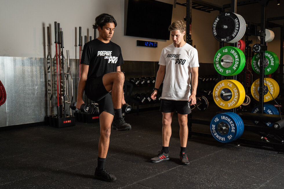 Two athletes in a gym, one lifting weights and the other observing, both wearing Prepare Like a Pro attire.