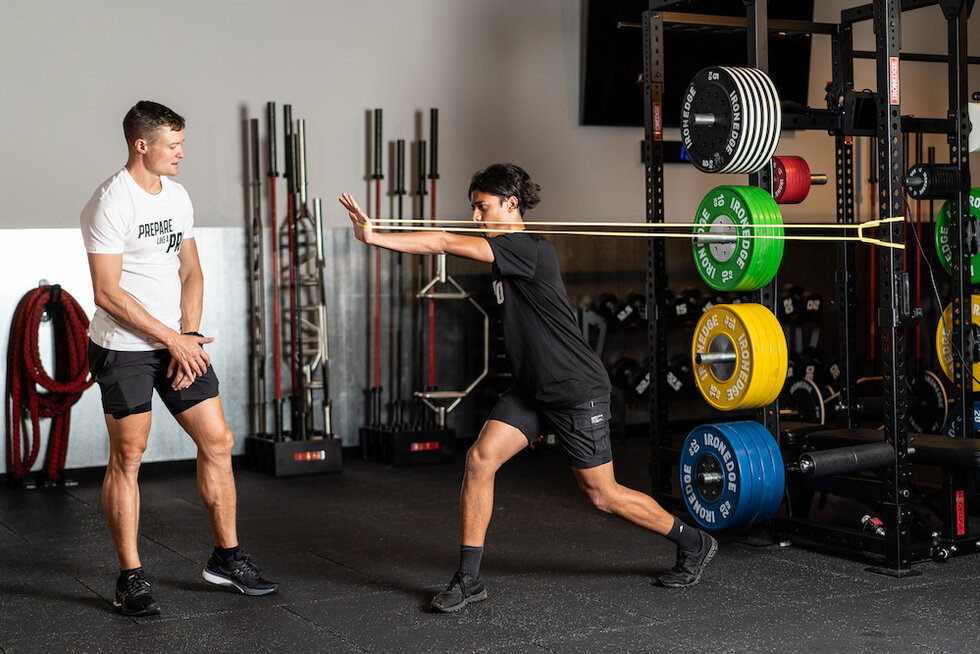 A trainer named Jack McLean observes a client performing a dynamic stretching exercise in a gym.