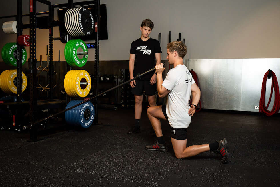 Two athletes in a gym, one performing a barbell lunge under the supervision of a trainer.