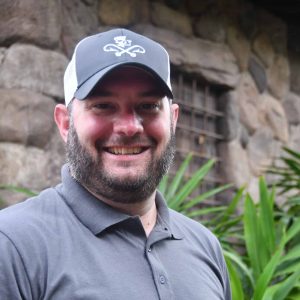 Friendly man with a beard wearing a gray polo shirt and black cap in a tropical garden setting.