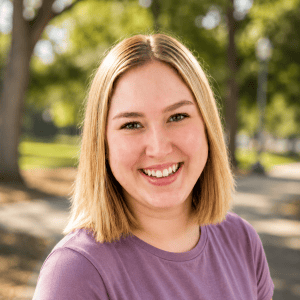 Smiling woman with blonde hair in a purple shirt outdoors in a park.