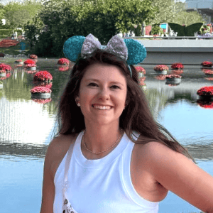 Disney-themed Minnie Mouse ears headband, smiling woman enjoying a park setting with a pond and flowers in the background.