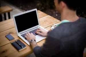 Man Typing On A Laptop At An Outdoor Wooden Table