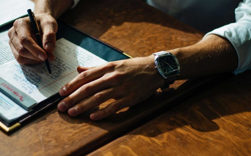 ALT text: Person writing notes at a wooden desk with a watch on their wrist.