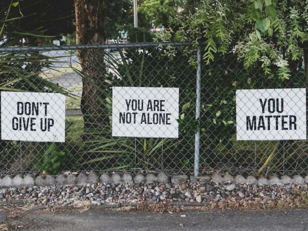 three signs on a fence saying, "DON'T GIVE UP," "YOU ARE NOT ALONE," and "YOU MATTER"