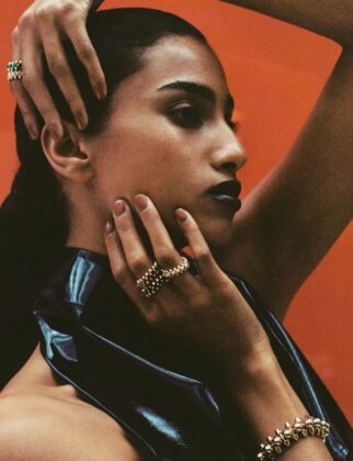 Imaan Hammam wearing a dark practical jacket, standing with her back to the camera, looking out over a still glacial lake reflecting a snow-capped mountain range at dawn.