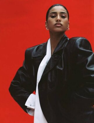 Imaan Hammam wearing a dark practical jacket, standing with her back to the camera, looking out over a still glacial lake reflecting a snow-capped mountain range at dawn.