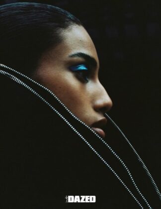 Imaan Hammam wearing a dark practical jacket, standing with her back to the camera, looking out over a still glacial lake reflecting a snow-capped mountain range at dawn.