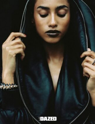 Imaan Hammam wearing a dark practical jacket, standing with her back to the camera, looking out over a still glacial lake reflecting a snow-capped mountain range at dawn.