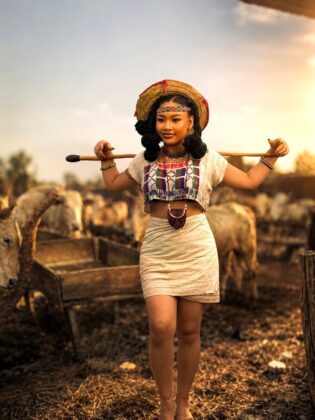Pearl Shim Mugalla posing in traditional Fulani attire with a camel in a dusty Sahel landscape