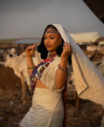 Pearl Shim Mugalla posing in traditional Fulani attire with a camel in a dusty Sahel landscape