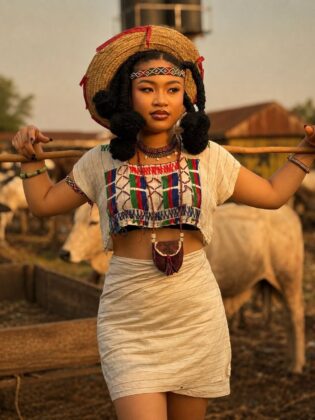 Pearl Shim Mugalla posing in traditional Fulani attire with a camel in a dusty Sahel landscape