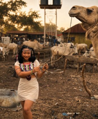 Pearl Shim Mugalla posing in traditional Fulani attire with a camel in a dusty Sahel landscape