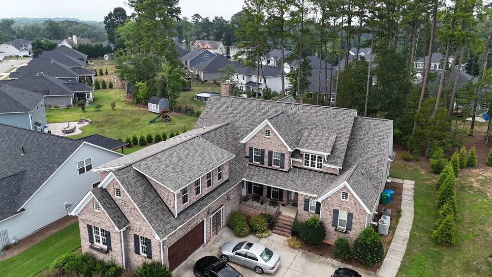 Drone view of a brick home with a new weathered-wood shingle roof in a wooded Charlotte neighborhood