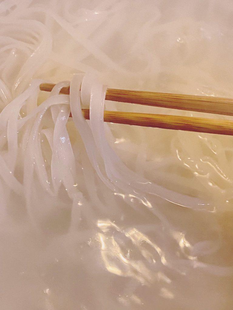 Cooked rice noodles being lifted with chopsticks from boiling water.
