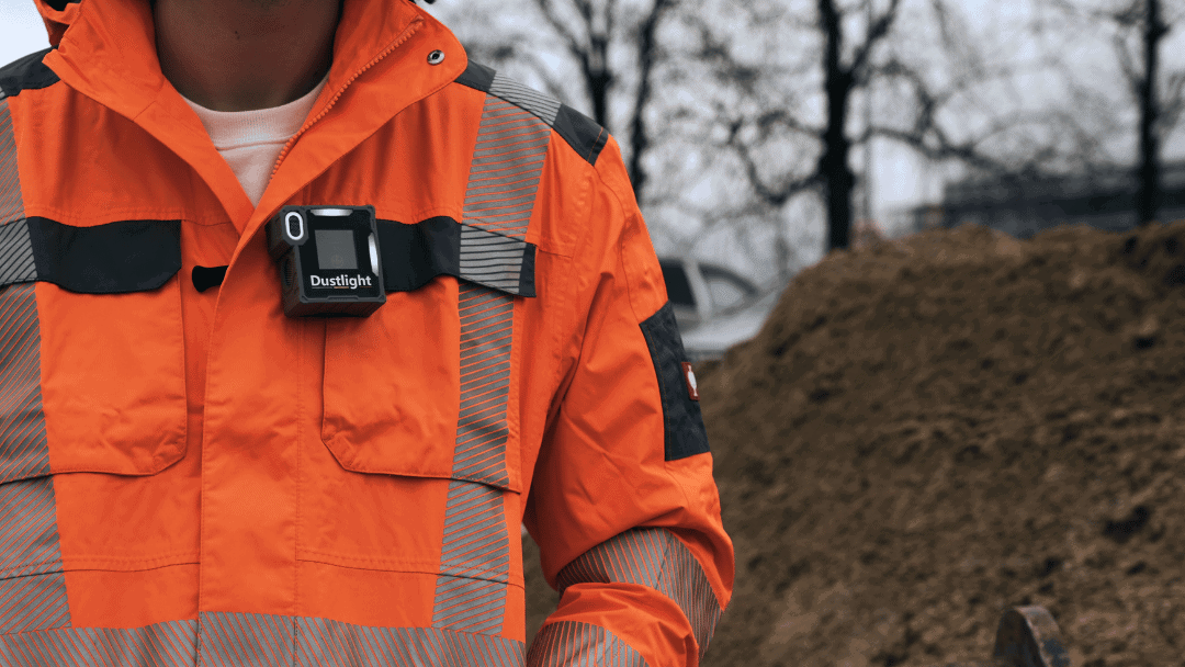 Arbeiter in orangefarbener Schutzkleidung trägt das tragbare Feinstaubmessgerät Dustlight zur Echtzeit-Überwachung von PM1, PM2.5 und PM10 auf einer Baustelle.