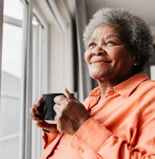 Senior woman holding mug, smiling and looking into distance beside window at home
