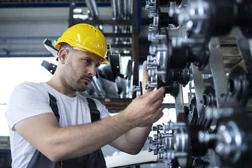 Maintenance technician in a yellow hard hat performing preventive maintenance on industrial conveyor rollers inside a factory.