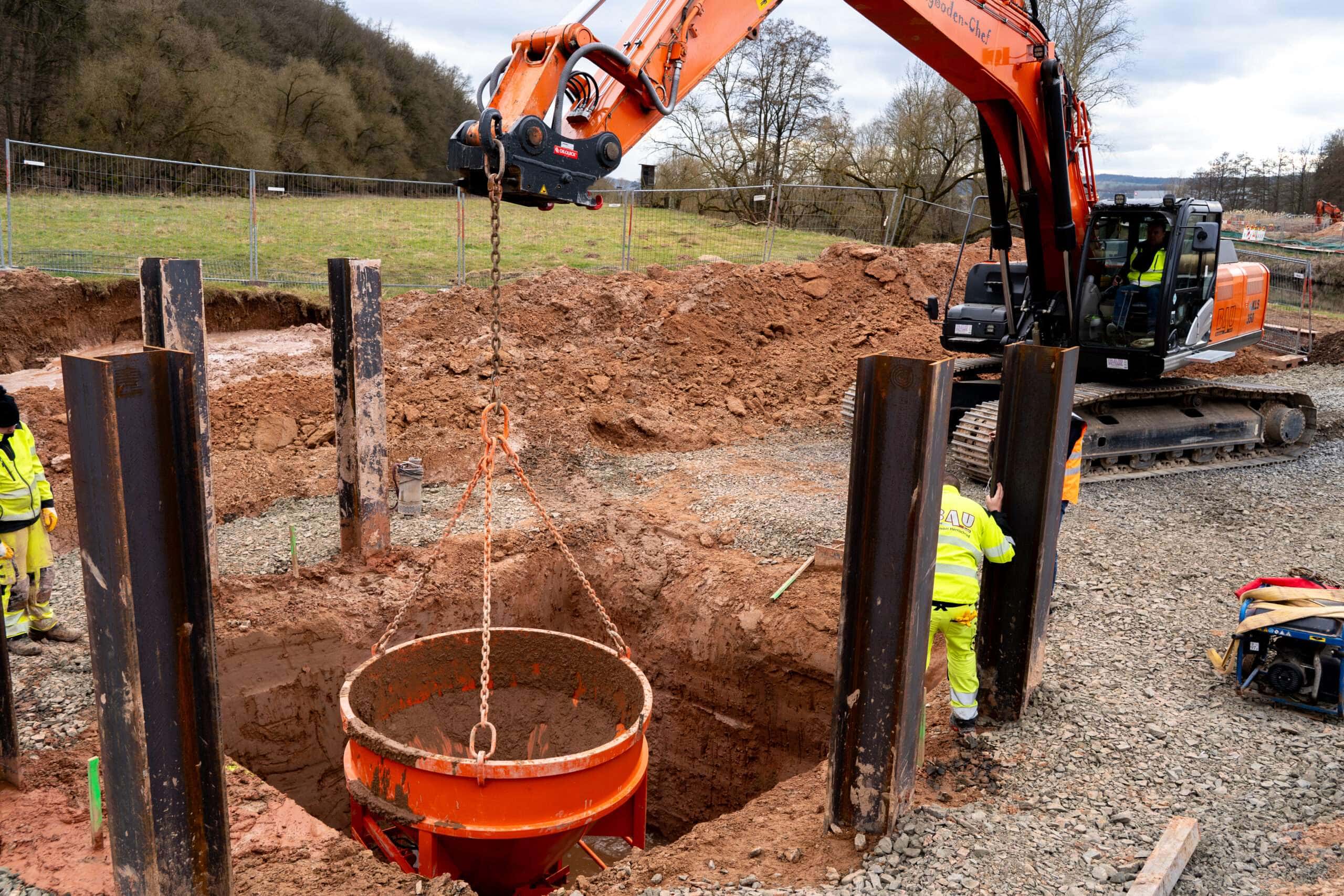 Ein Baustellenfahrzeug lässt einen Metalltrichter in die Baugrube herab, die nun mit RSS Flüssigboden verfüllt wird.