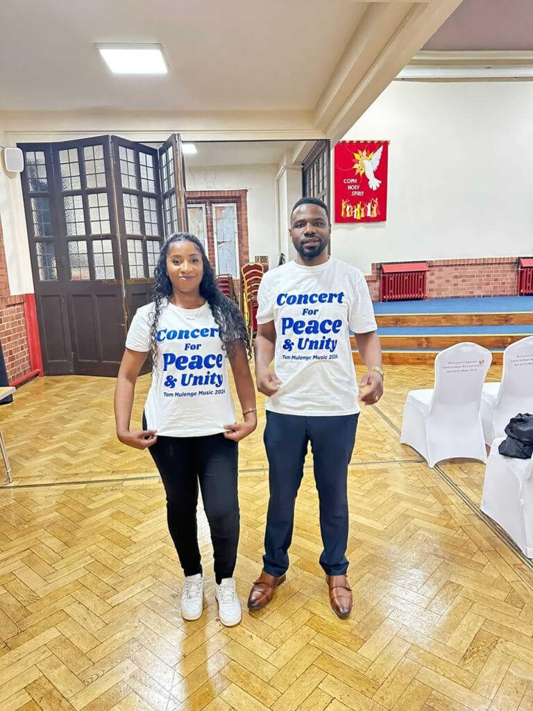 Wide editorial photograph of Tom Mulenge against a light backdrop, concert for peace and unity clothing.