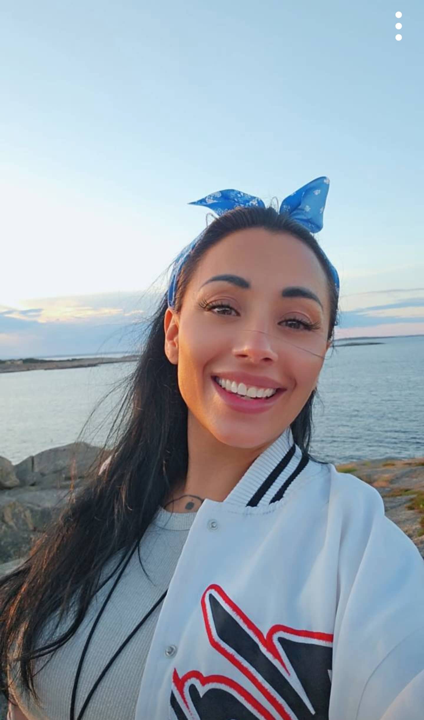 CatalayaX standing near calm water at sunset wearing a blue bandana and white jacket.