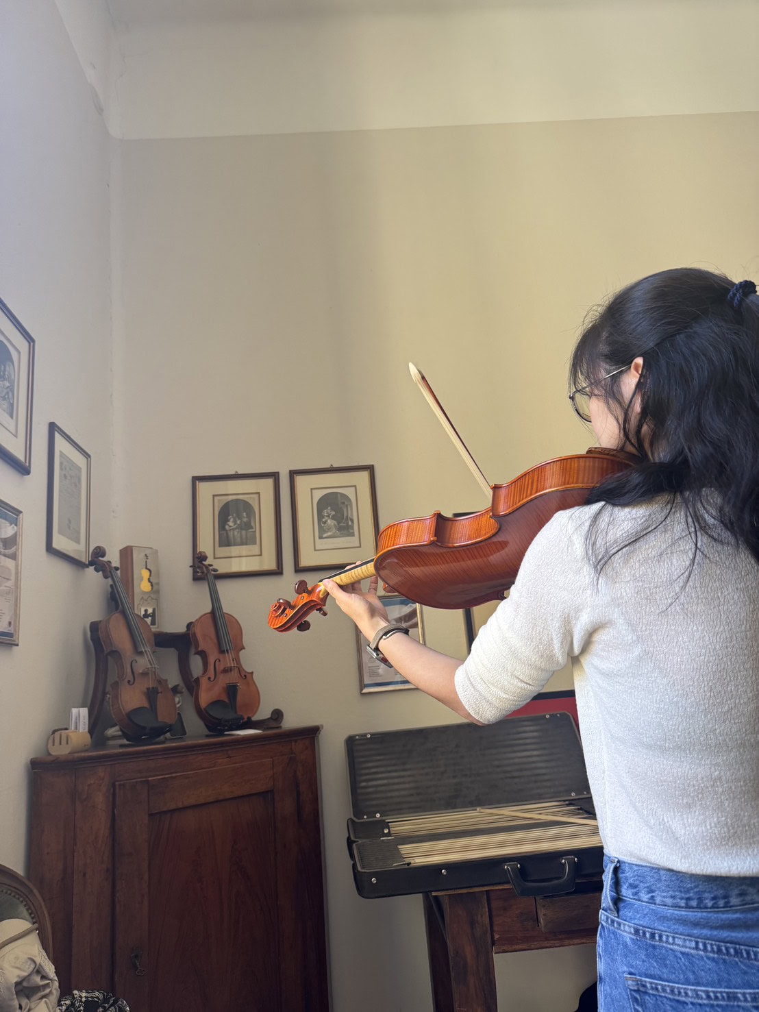 Person playing a wooden violin in a cozy room, standing at a music stand with a violin case nearby.