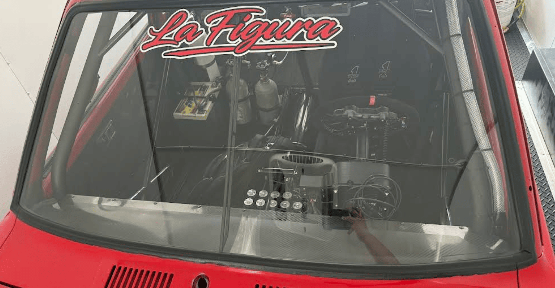 Cockpit of a race car viewed through the windshield, showing a racing steering wheel, roll cage, gauges, and the 'La Figura' decal on the glass.