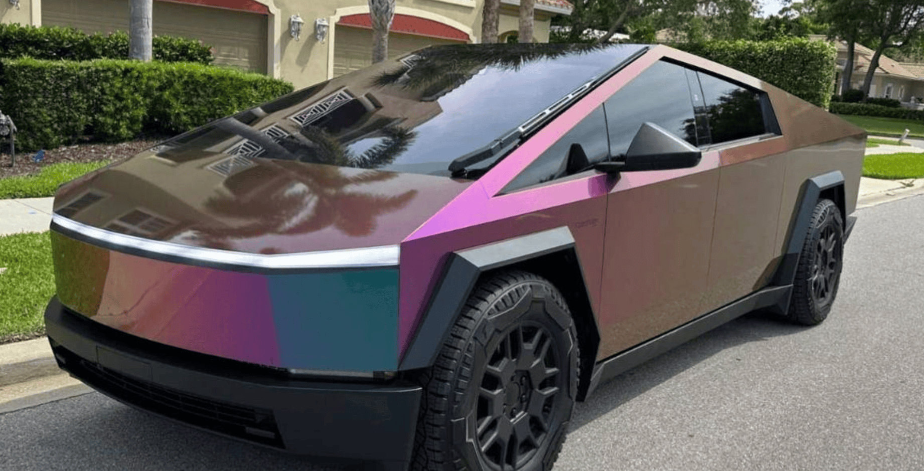 Colorful, angular-wrapped electric SUV (Tesla Cybertruck) parked on a suburban street, showcasing a multicolor geometric wrap with pink, purple, and brown panels.