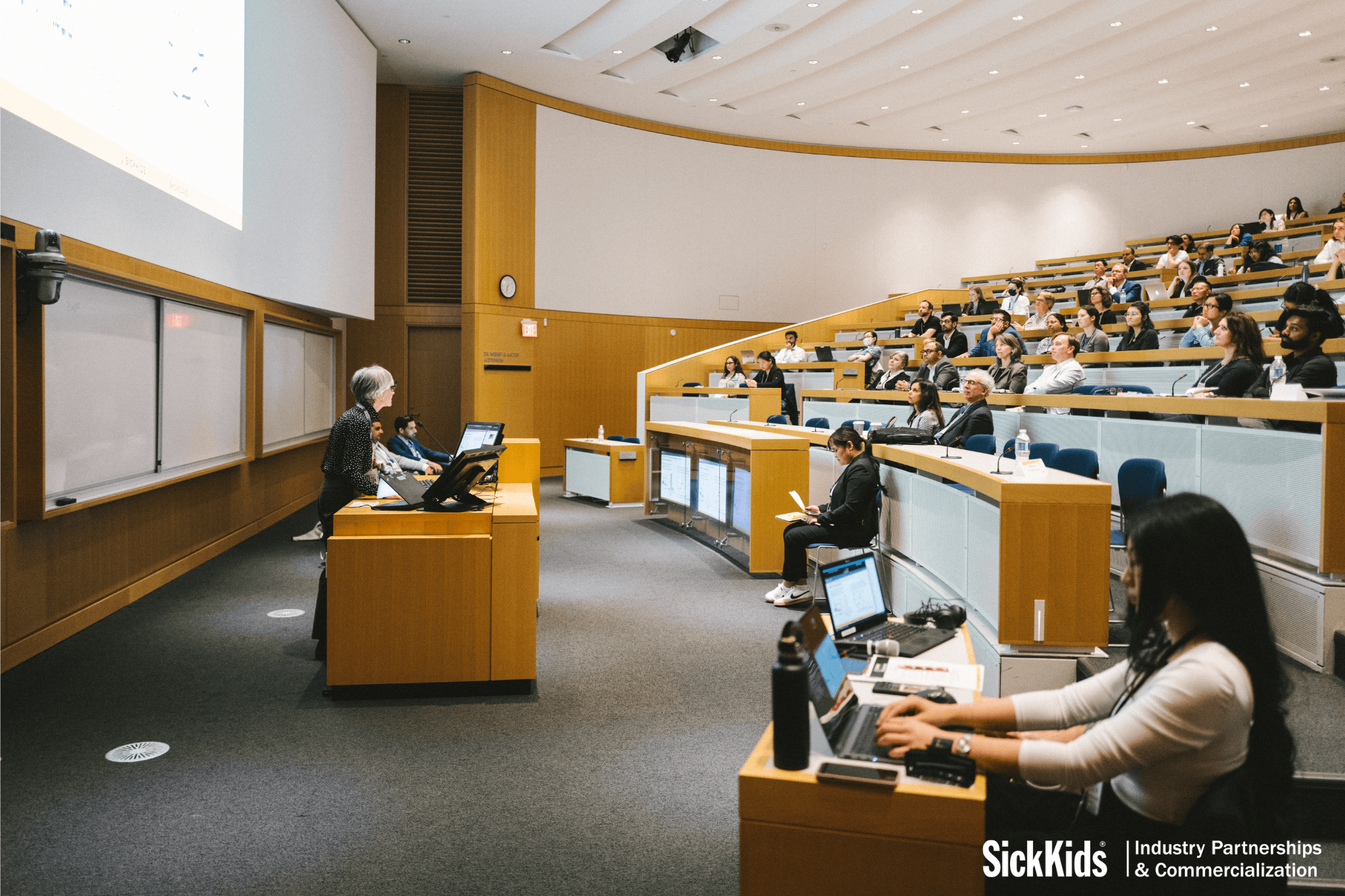 Crowd of industry partners sitting in auditorium while a SickKids researcher pitches their project at the SIckKids Innovation Showcase
