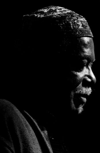 Profile of an older man with a patterned headwrap against a solid black background.