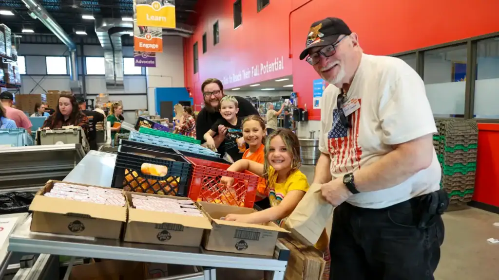 Two adults and three children standing at a Sack Supper packing table pause to smile.