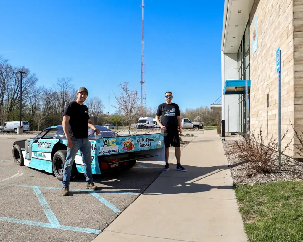 Adam Casari and another adult smile and stand at the bumper of his race car, which includes the Kids' Food Basket logo.
