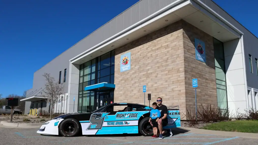 Adam Casari poses for a photo with his son in front of his blue, white and silver race car parked in front of Kids' Food Basket.