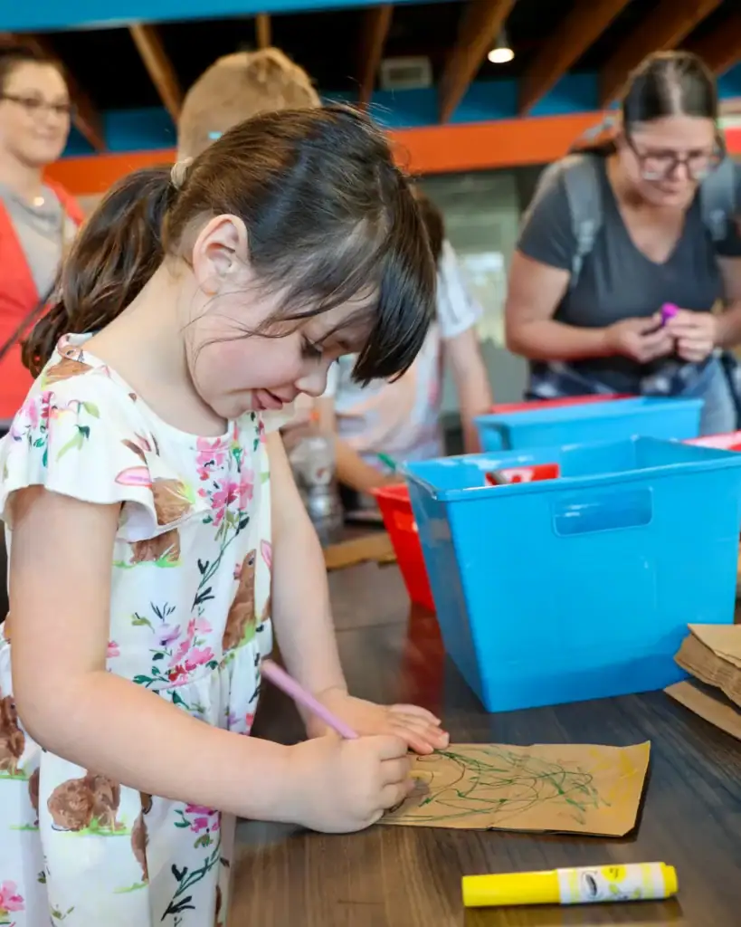 A girl bent over a table colors a brown paper bag.