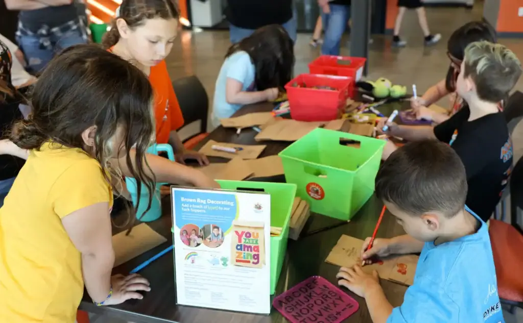Children seated at a table draw and color brown paper bags.