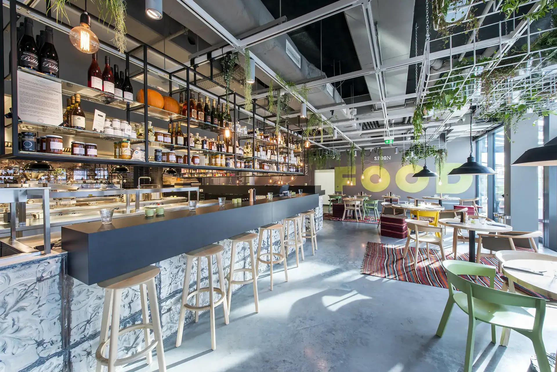 Blick in den Gastraum des Restaurant "Dachgarten" in Mannheim. Mittig sieht man die Bar, Tresen und Barhocker. Darüber hängend von der Decke einige Regale. Auf der rechten Bildseite stehen einige Sitzgruppen, bestehend aus Tischen und farbenfrohen Stühlen. An der Rückwand ist in großen Buchstaben das Word FOOD dargestellt.