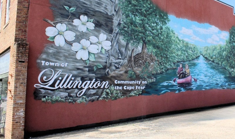 Mural on a red brick wall reading 'Town of Lillington' with a river scene, two people in a canoe on Cape Fear, and white dogwood blossoms.